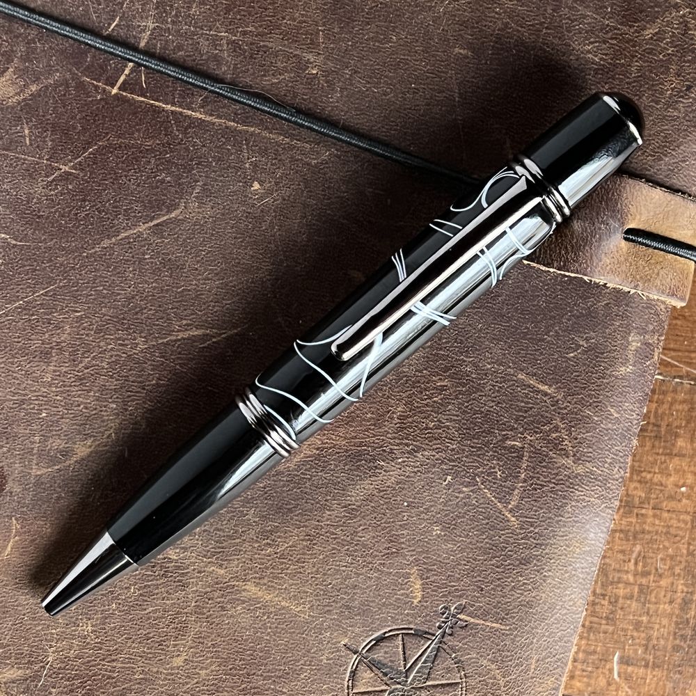 A thick bodied twist pen in dark chrome and black and white colors on a leather journal